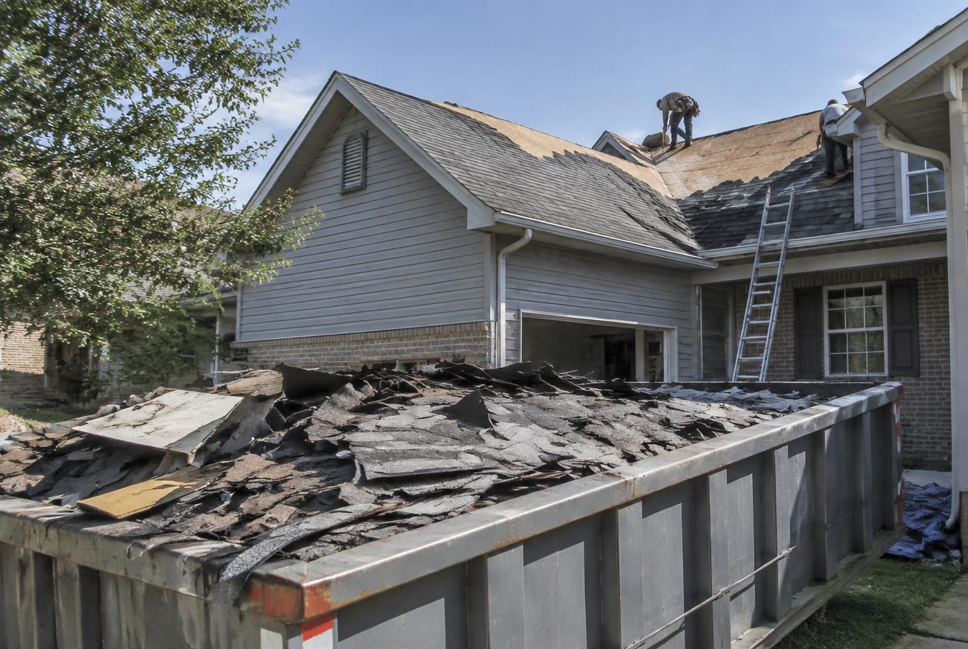Torn-off asphalt shingles filling a roll-off dumpster beside a house during a roof replacement